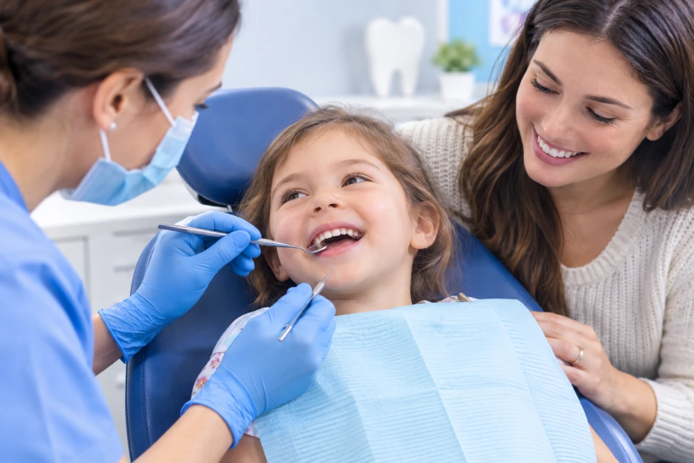 Child sits in dental chair smiling as dentist uses mirror and dental tools; mother watches supportively.
